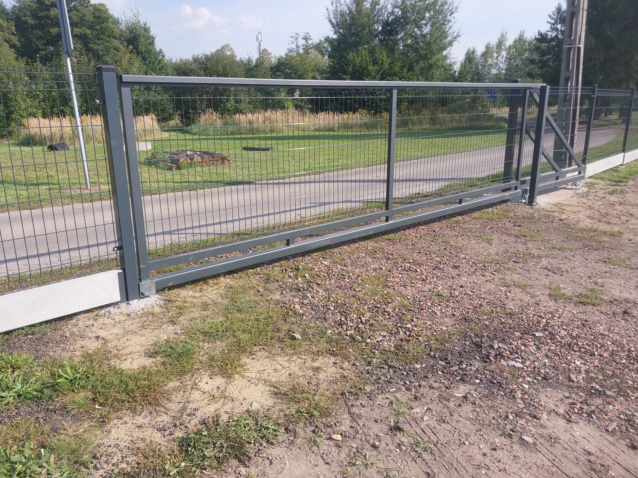 Przesuwna brama z siatki metalowej w kolorze szarym, zamontowana na betonowej podmurówce, widok z zewnątrz na tle zieleni i drogi.
