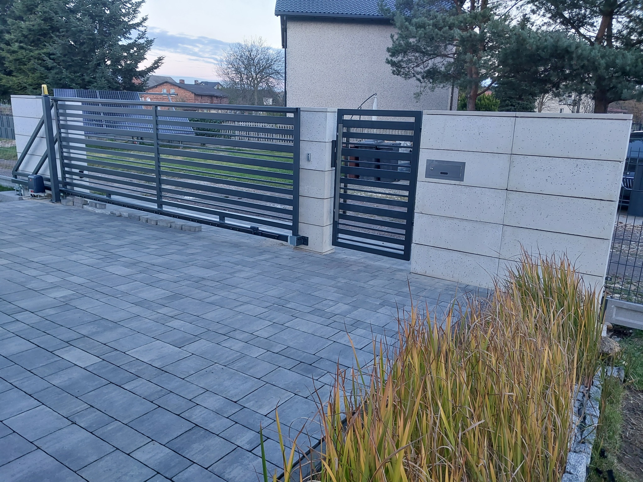 Nowoczesna, przesuwna brama metalowa z poziomymi szczeblami, zamontowana na betonowych słupkach, z widocznym automatem do bramy i kostką brukową na podjeździe, otoczona trawą ozdobną.