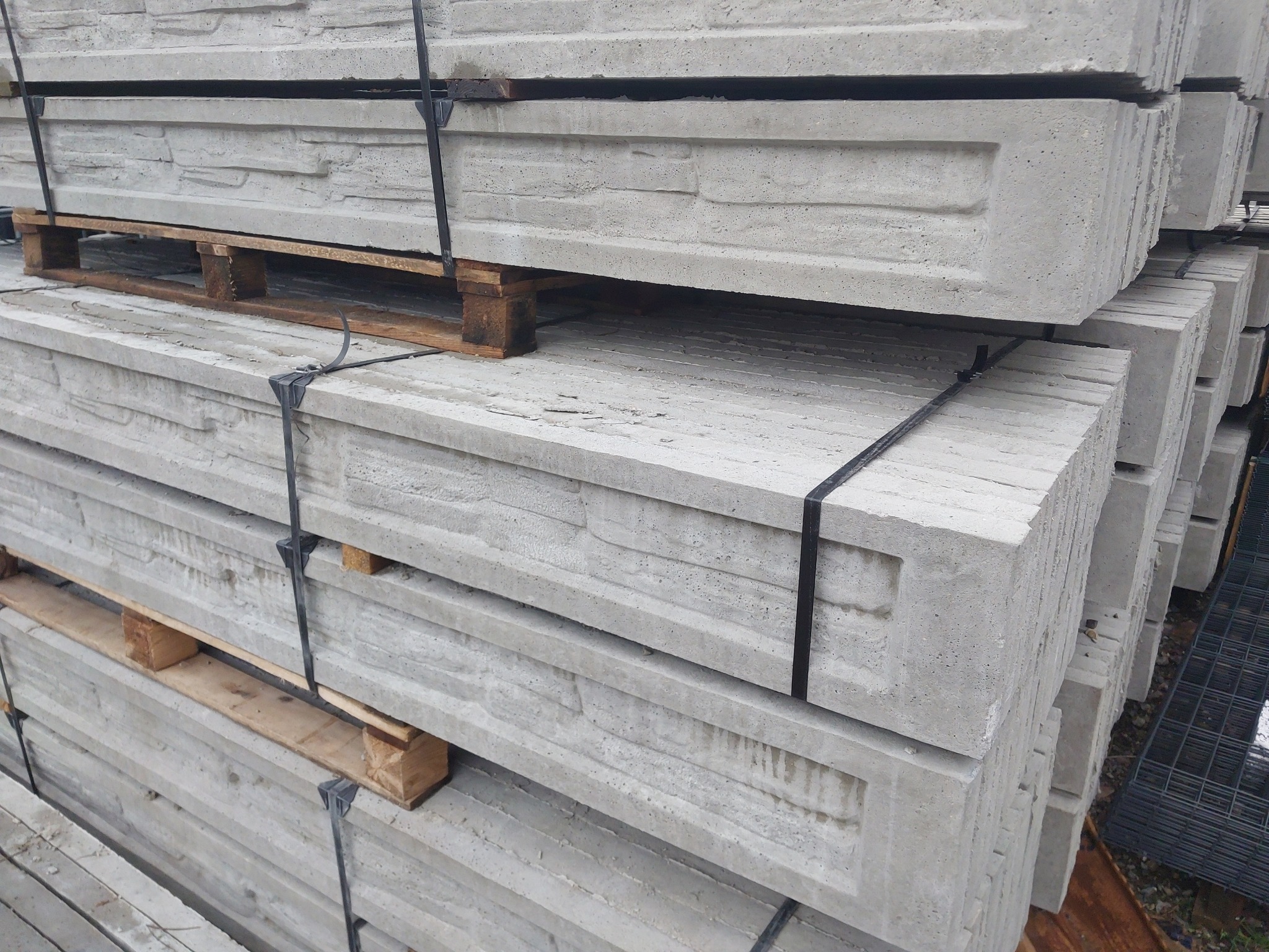 Stos betonowych prefabrykatów imitujących kamień, spiętych czarną taśmą na palecie, przeznaczonych do budowy ogrodzenia.