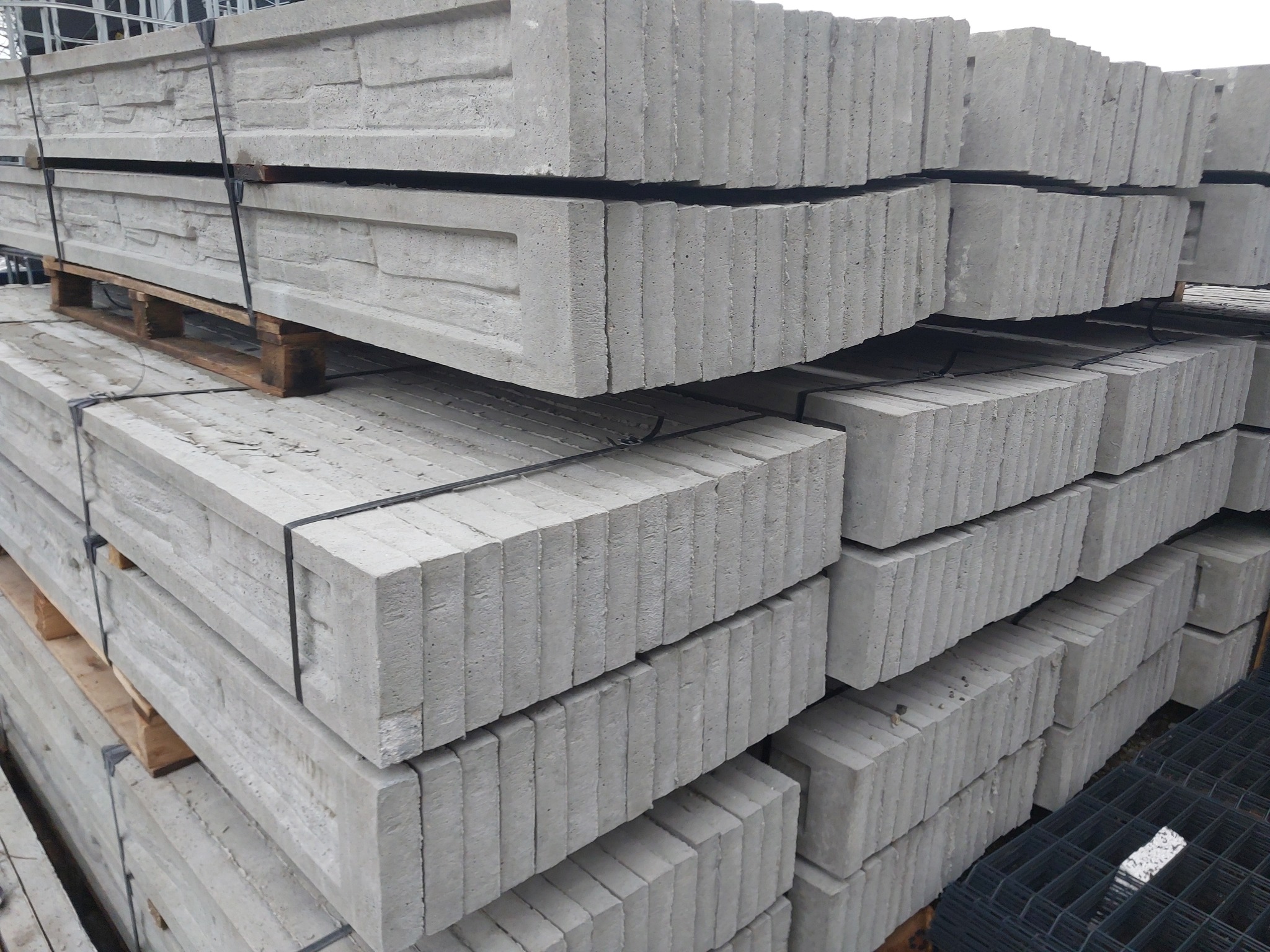 Stosy prefabrykowanych, betonowych elementów ogrodzeniowych imitujących kamień, ułożonych na paletach i zabezpieczonych taśmą.