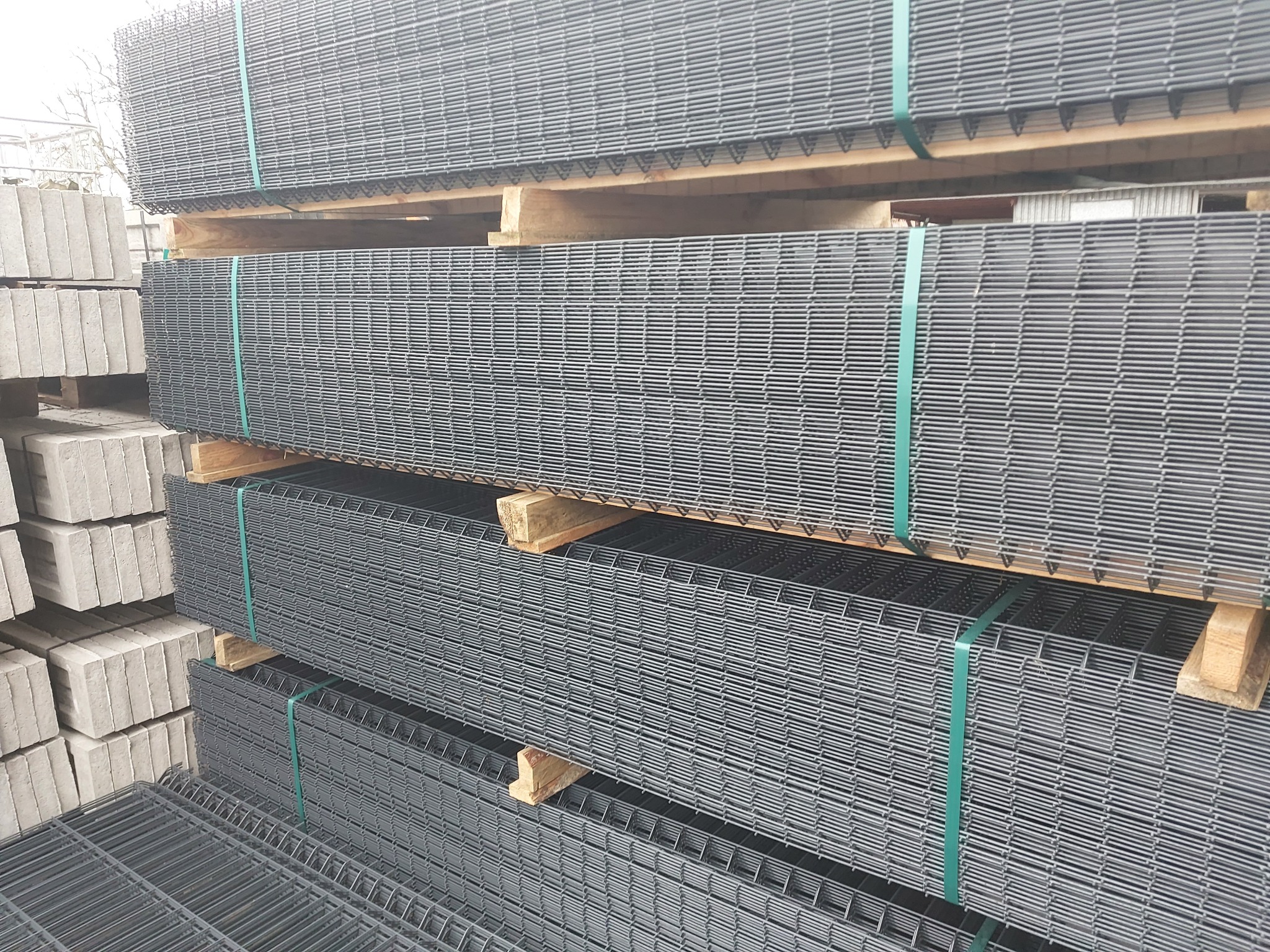 Stosy paneli ogrodzeniowych zgrzewanych, zabezpieczonych zieloną taśmą, ułożone na drewnianych paletach, obok stosu betonowych bloczków.