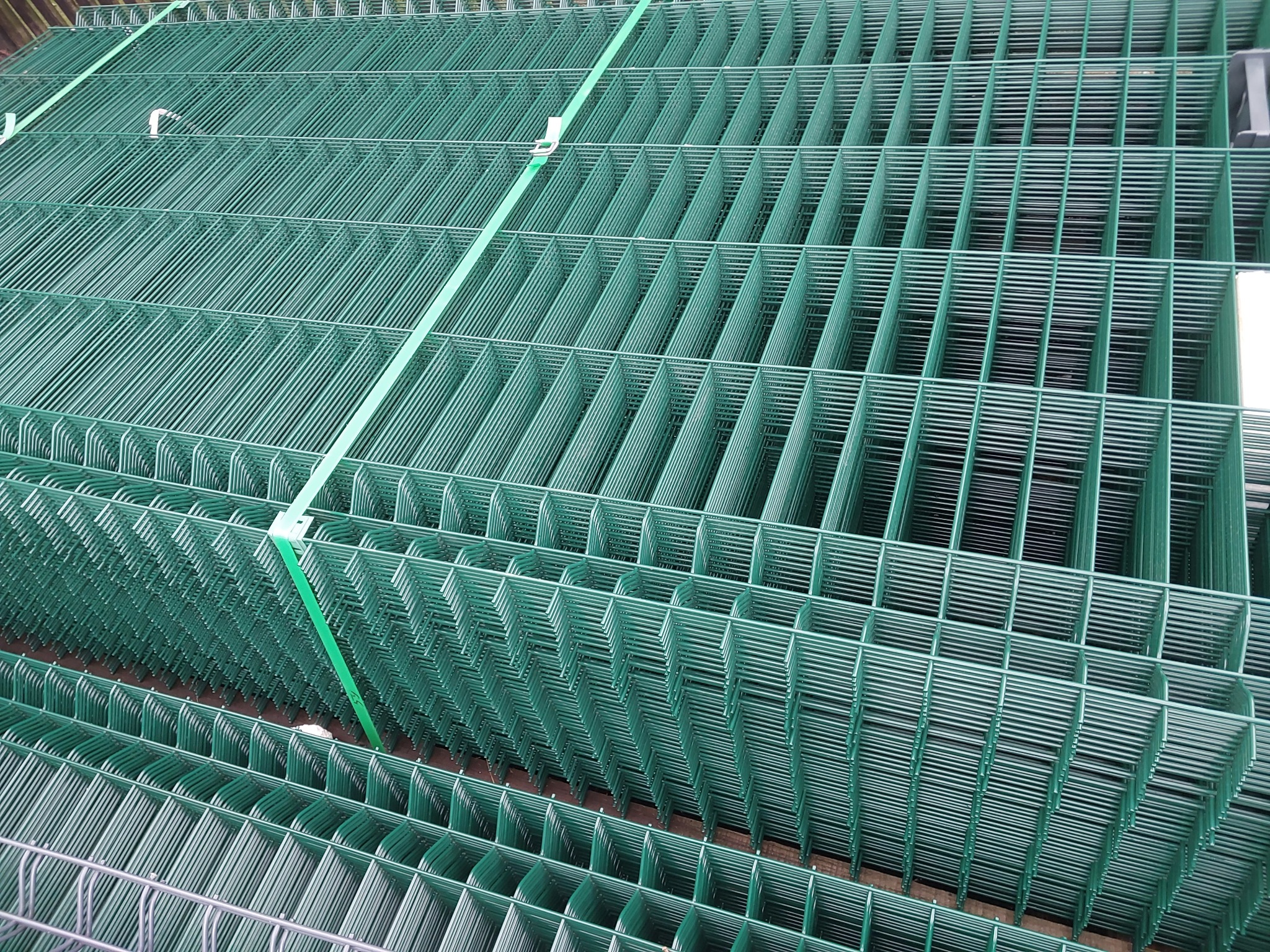 Stos zielonych, metalowych paneli ogrodzeniowych zabezpieczonych zieloną taśmą transportową.