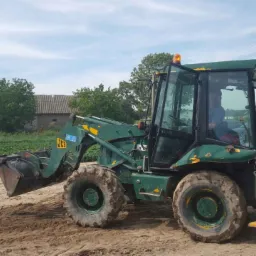 Zielona koparko-ładowarka JCB na placu budowy, widok z boku, zarys budynku w tle, lekko pochmurne niebo. Maszyna z widocznymi śladami użytkowania.
