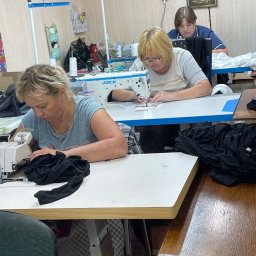 Stanitska - Praca w szwalni: trzy szwaczki przy maszynach Jack, szyjące czarne i białe tkaniny. Jasne wnętrze, drobne akcesoria na stołach, kalendarz na ścianie.