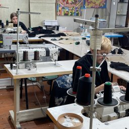 Stanitska - Praca w szwalni: dwie krawcowe przy maszynach, czarne nici na szpulach, jasne blaty stołów, oświetlenie LED, kolorowe obrazy na ścianie w tle.