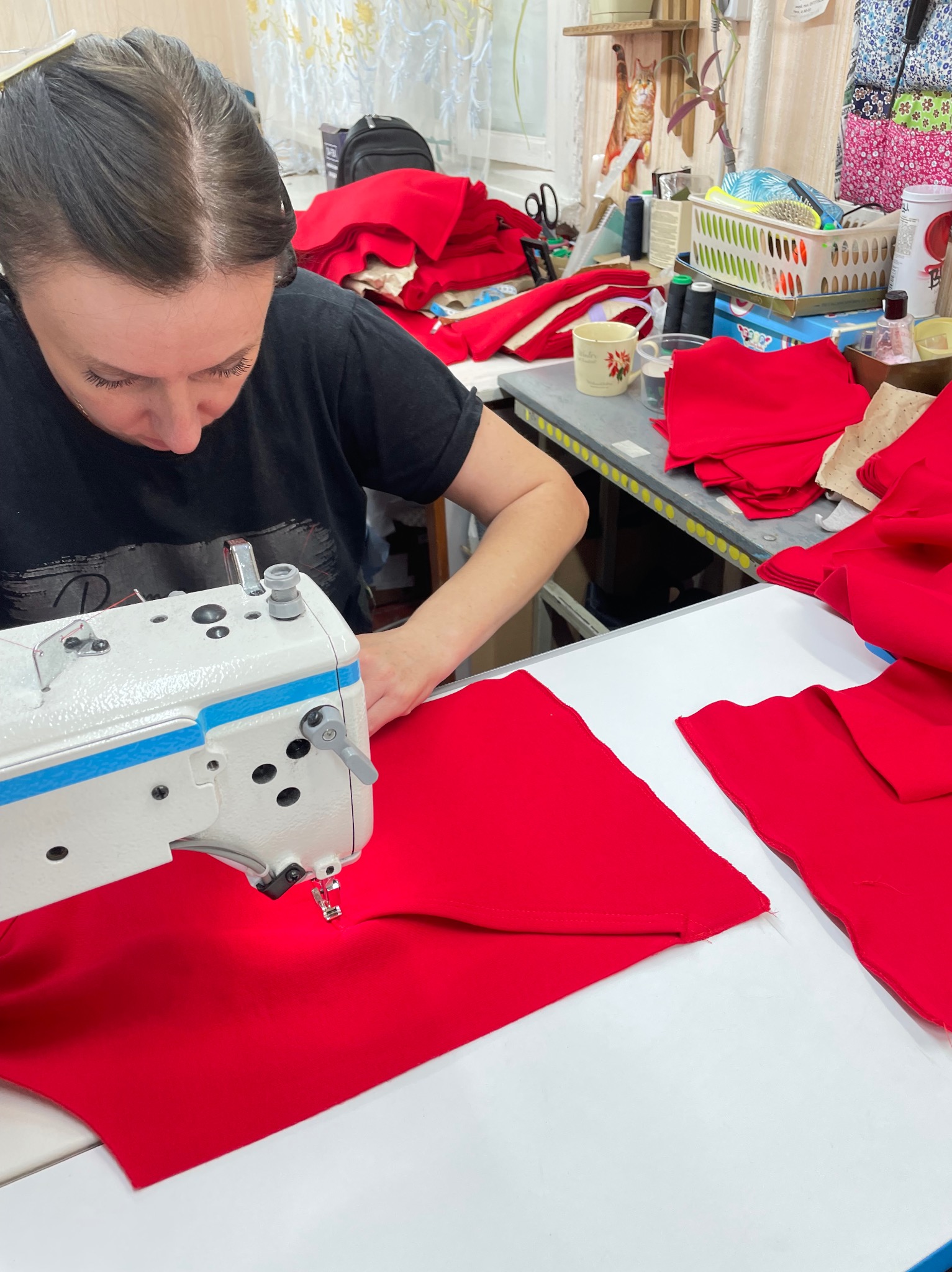 Kobieta szyje na maszynie przemysłowej czerwoną tkaninę. W tle widoczne stosy materiałów i akcesoria krawieckie na stole.