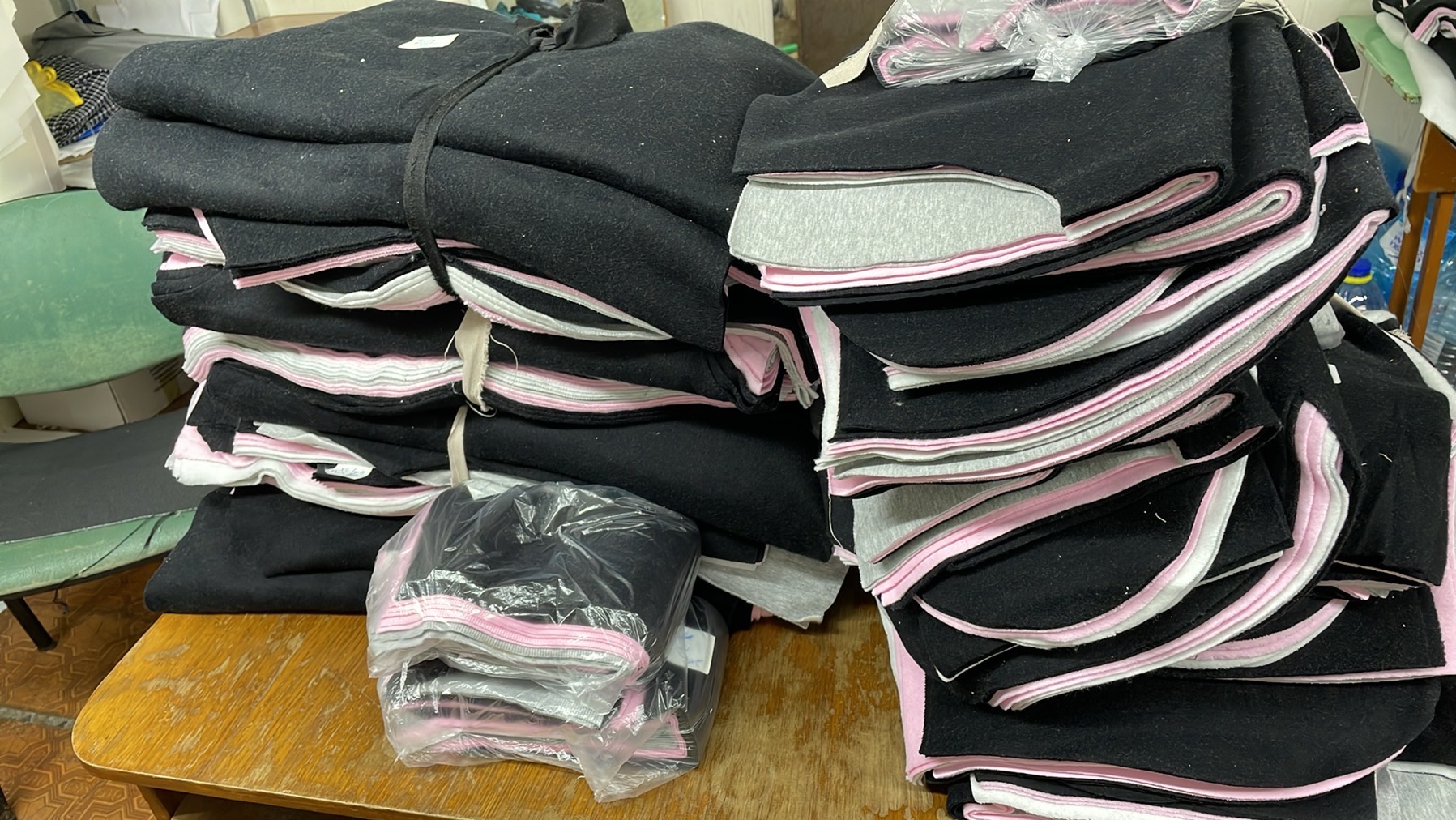 Stos czarnych, wykrojonych elementów odzieży z różowymi i szarymi podszewkami, ułożonych na drewnianym stole w szwalni. Część spakowana w folię.