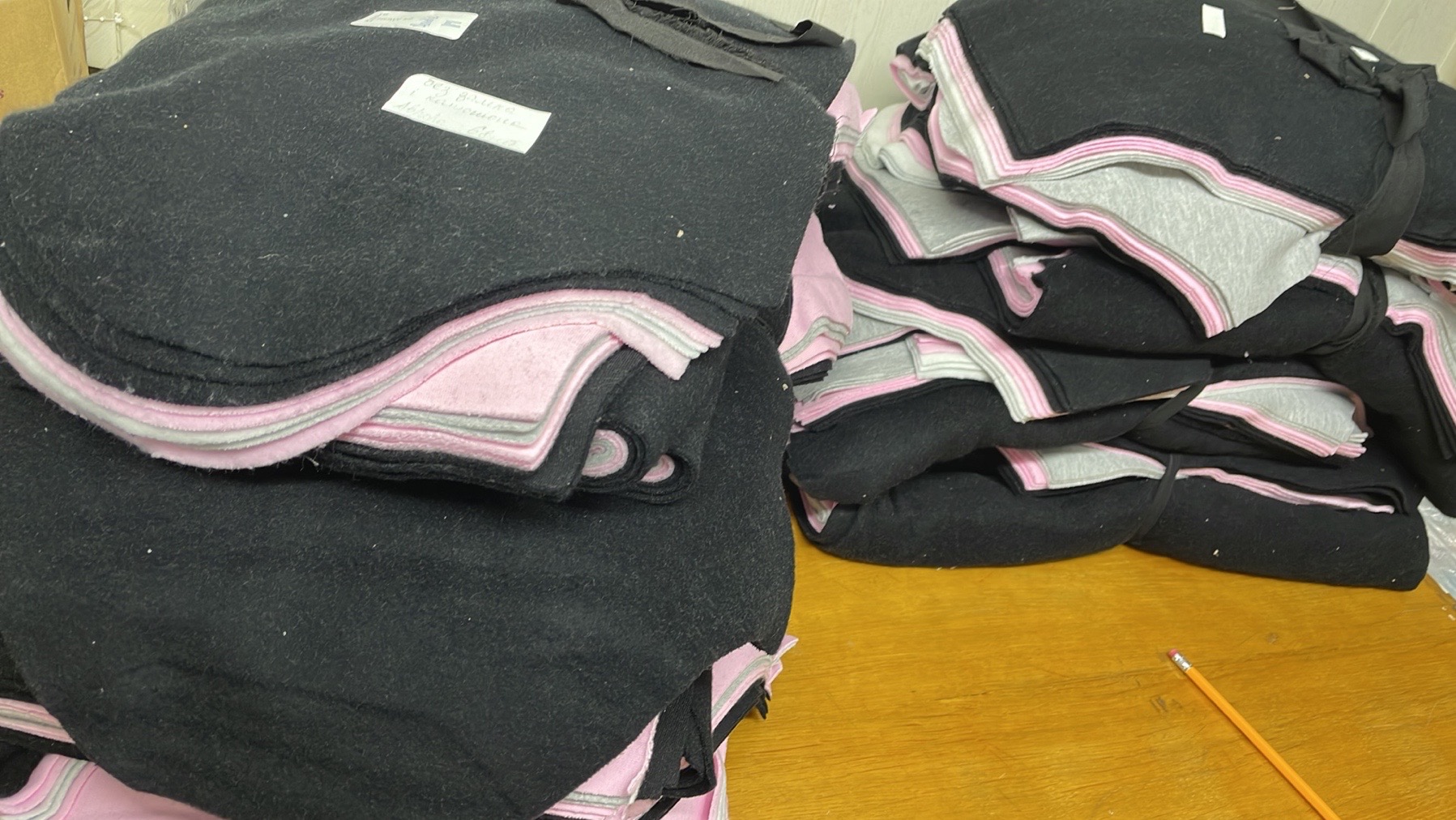 Stos czarnych, miękkich kocyków z różowo-szarym obszyciem, ułożonych na drewnianym stole. Na jednym z kocyków widoczna biała kartka z napisem.