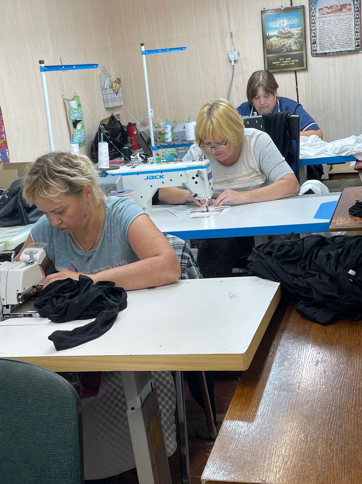 Praca w szwalni: trzy szwaczki przy maszynach Jack, szyjące czarne i białe tkaniny. Jasne wnętrze, drobne akcesoria na stołach, kalendarz na ścianie.