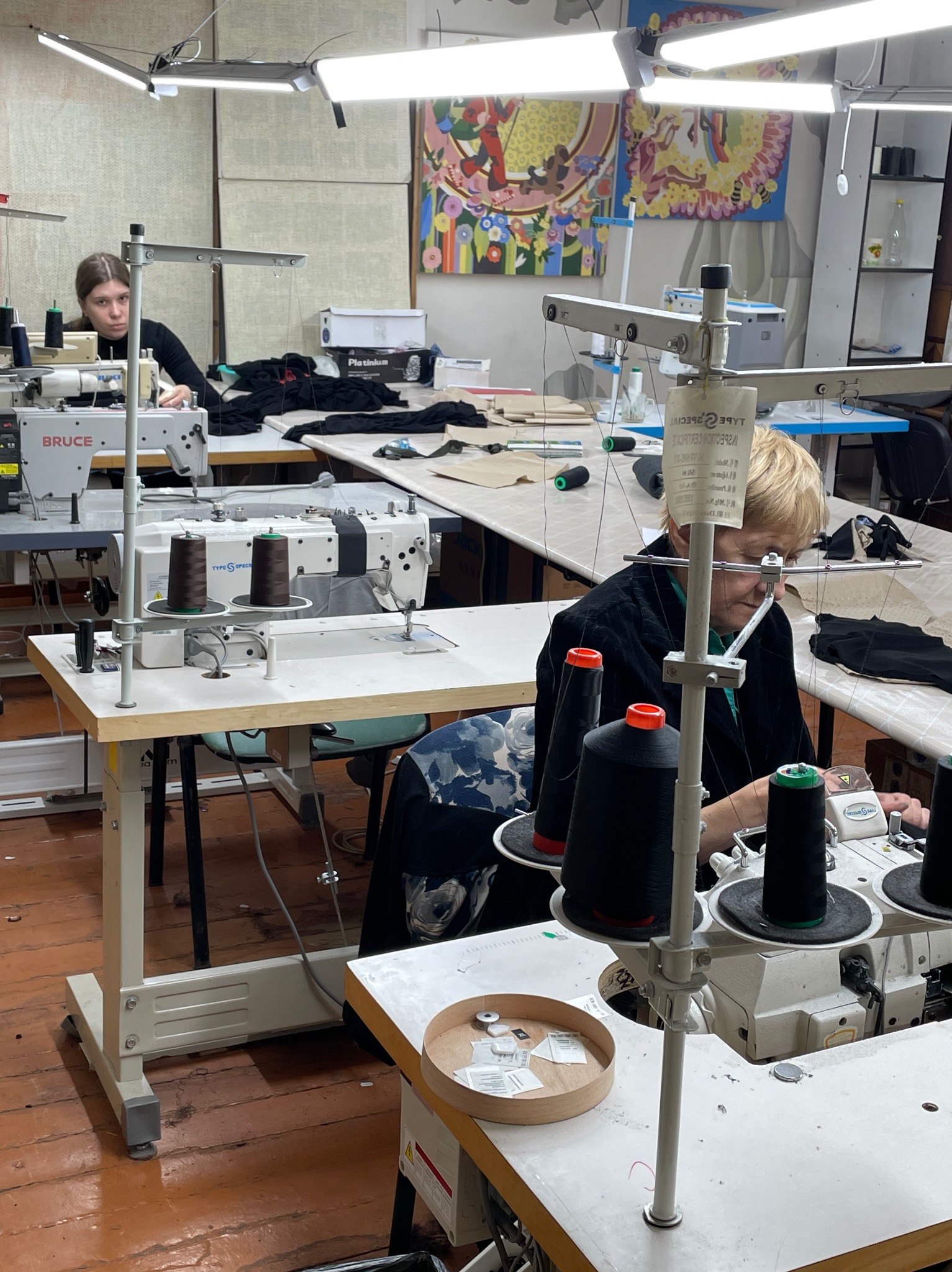 Praca w szwalni: dwie krawcowe przy maszynach, czarne nici na szpulach, jasne blaty stołów, oświetlenie LED, kolorowe obrazy na ścianie w tle.