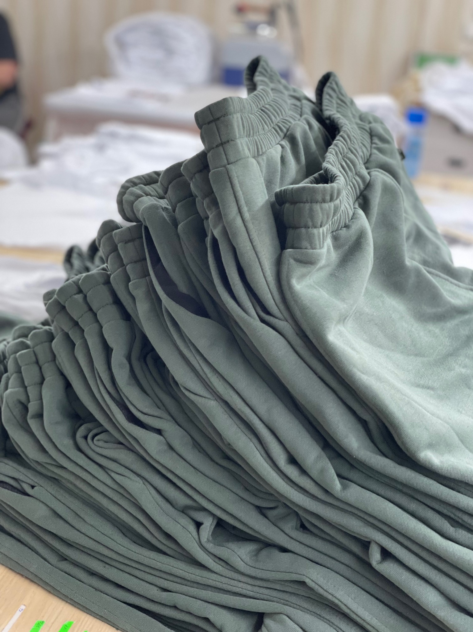 Stos uszytych, zielonych spodni dresowych na stole w szwalni. Widoczny elastyczny pas i ściągacze. W tle rozmyte białe tkaniny i maszyny.