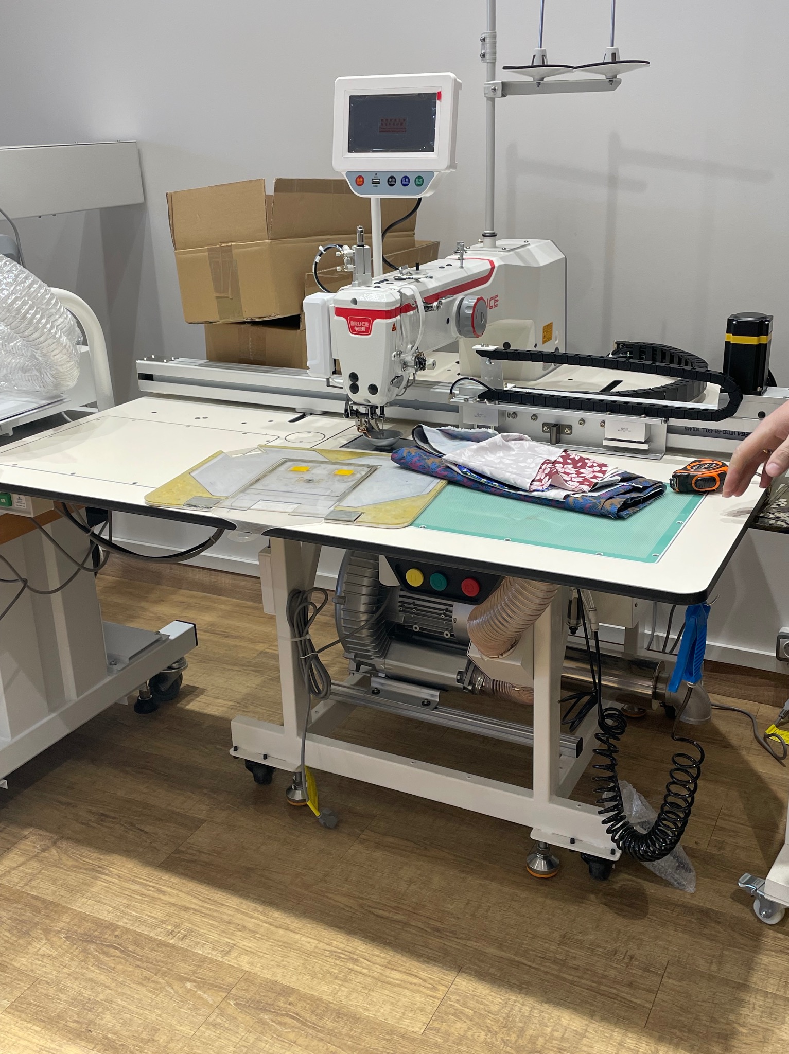 Automat szwalniczy Bruce na stole z materiałami i miarką, gotowy do pracy. W tle kartony i elementy wyposażenia szwalni.