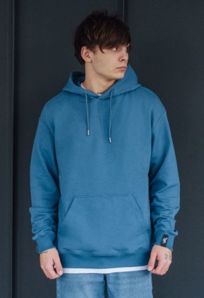 Młody mężczyzna prezentuje błękitną bluzę z kapturem, widoczne logo na mankiecie, jasne jeansy i ciemnoszare tło.