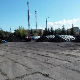 Składowisko węgla z hałdami przykrytymi plandekami, widoczny czerwony ciągnik i wysokie maszty telekomunikacyjne w tle.