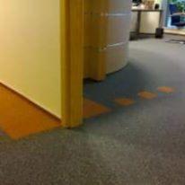 Fragment biura z nową wykładziną dywanową w odcieniach szarości i pomarańczy, widoczny narożnik ściany i fragment recepcji w tle.