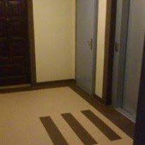 Beżowa podłoga z ciemnobrązowymi, równoległymi paskami, widoczna w korytarzu z ciemnymi drzwiami po lewej i szarymi drzwiami po prawej stronie. Ujęcie z niskiego kąta, podkreślające wzór podłogi.