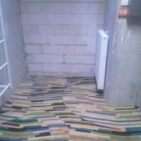 Fragment pomieszczenia w trakcie prac wykończeniowych, widoczne niedokładnie ułożone, kolorowe deski podłogowe na nierównym podłożu, surowe ściany z betonu komórkowego i biały grzejnik.