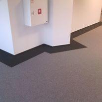 Szara tekstylna powierzchnia podłogowa z ciemnoszarymi listwami przypodłogowymi w narożniku pomieszczenia z białymi ścianami i skrzynką alarmową.