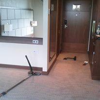 Wnętrze pokoju hotelowego w trakcie odświeżania. Widoczny odkurzacz piorący na beżowej wykładzinie dywanowej, fragment ściany z imitacją cegieł oraz ciemne drzwi.