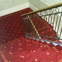 Czerwona, wzorzysta powłoka schodowa z bordiurą, zamontowana na schodach z metalową balustradą i drewnianym poręczą.