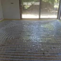 Białe rury ogrzewania podłogowego ułożone na srebrnej folii, zamocowane do czarnej siatki na podłodze w jasnym pokoju z oknami na zewnątrz.