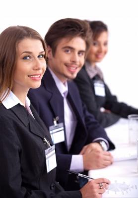 Trzy osoby w garniturach siedzą przy stole z identyfikatorami, uśmiechają się do kamery. Pierwszy plan ostry, tło rozmyte. Jasne, neutralne tło.