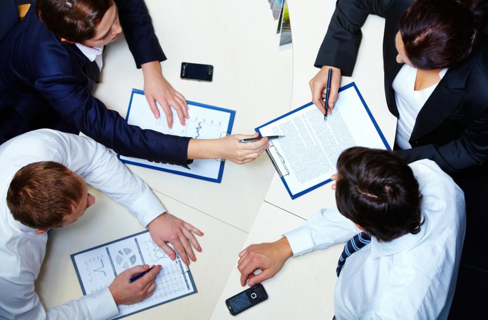 Zespół analizuje dokumenty i wykresy przy stole konferencyjnym, widok z góry. Smartfony leżą obok, podkreślając nowoczesny charakter spotkania.