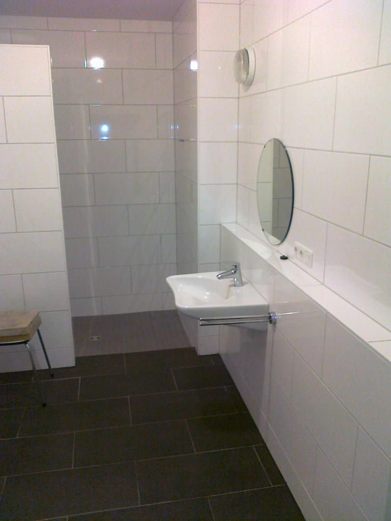 Nowoczesna łazienka z białą glazurą ścienną i ciemną podłogą. Umywalka wisząca, okrągłe lustro i minimalistyczny design.