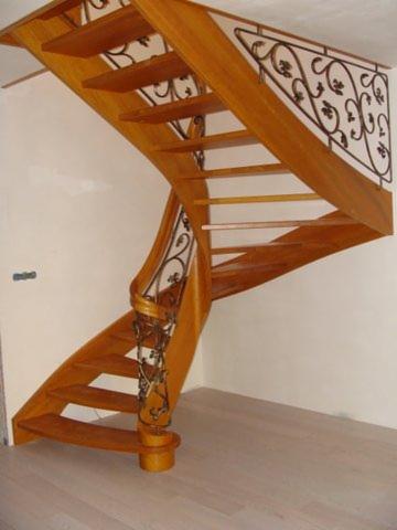 Drewniane schody spiralne z ozdobną, kutą balustradą, oświetlone naturalnym światłem. Parkiet na podłodze, jasne ściany. Widok z dołu, akcent na detale balustrady.