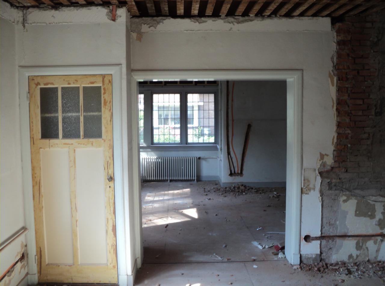Wnętrze w trakcie remontu: stara, drewniana, częściowo przeszklona, żółtawa stolarka drzwiowa z widocznymi śladami użytkowania, odsłonięta cegła na ścianie, fragmenty instalacji elektrycznej...