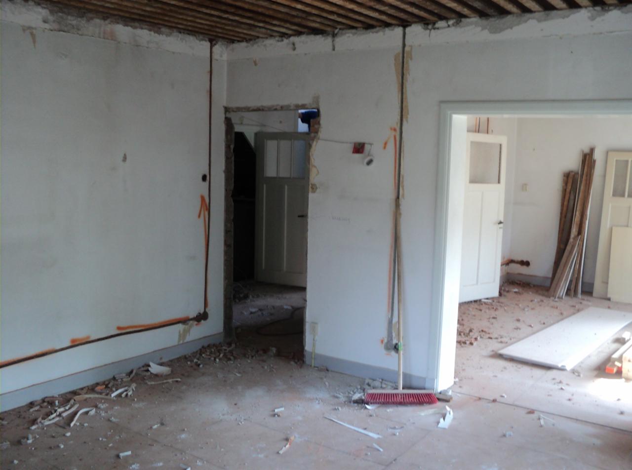 Wnętrze w trakcie remontu: odsłonięta instalacja elektryczna, fragmenty gruzu na podłodze, widoczne drzwi w przejściach. Czerwona zmiotka oparta o ścianę. Surowy stan.