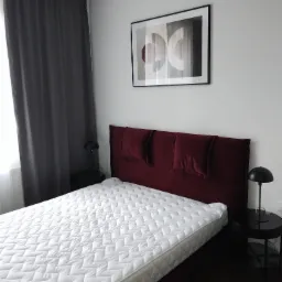 Sypialnia z łóżkiem z bordowym zagłówkiem i białą pikowaną narzutą, czarne stoliki nocne z lampkami, szare zasłony i abstrakcyjny obraz na ścianie.