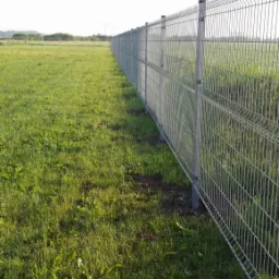 Długi, szary płot panelowy z metalowej siatki rozciąga się wzdłuż zielonego pola, w tle widoczne turbiny wiatrowe.