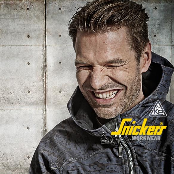 Uśmiechnięty mężczyzna w kurtce roboczej Snickers Workwear na tle betonowej ściany.