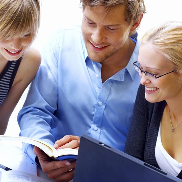 Trzy osoby (mężczyzna i dwie kobiety) pochylone nad laptopem i otwartą książką, uśmiechnięte, prawdopodobnie w trakcie nauki lub pracy, jasne tło.