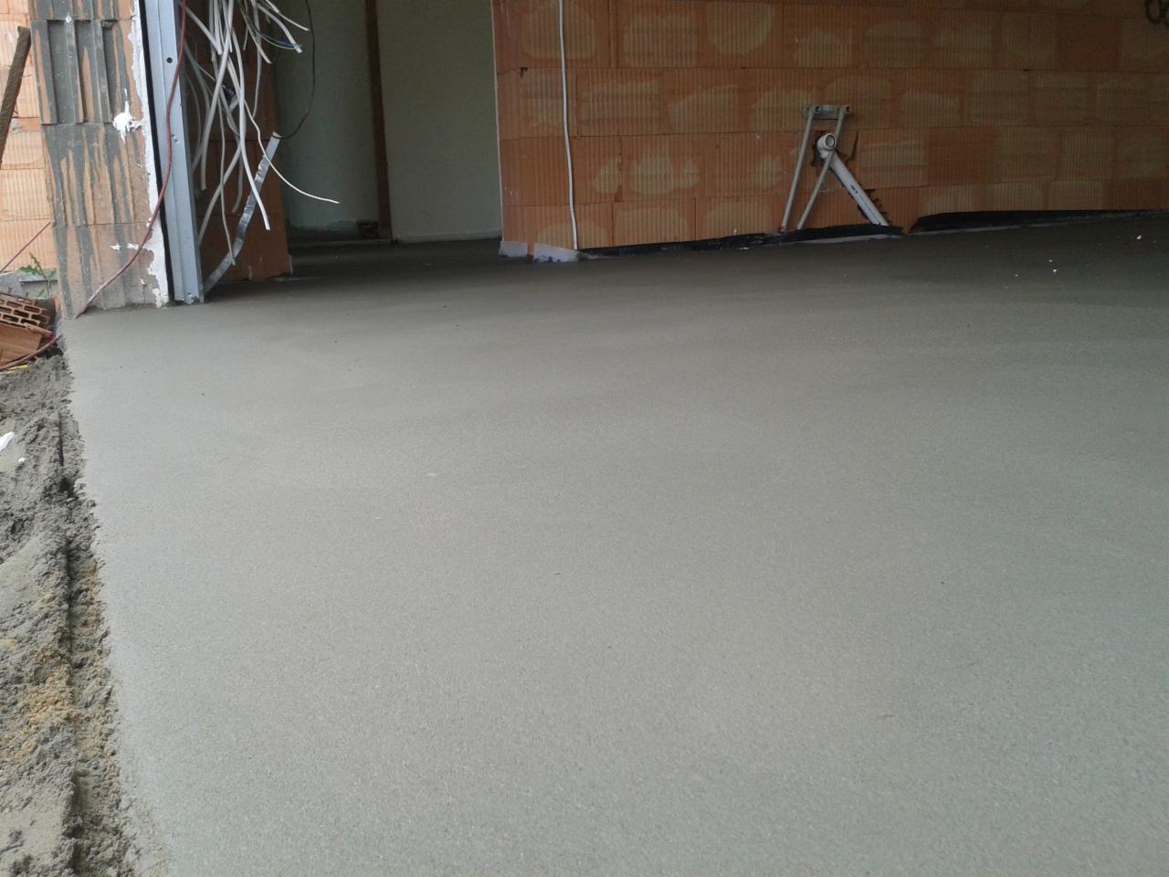 Świeżo wylana, gładka posadzka w surowym wnętrzu budynku z ceglanymi ścianami i widocznymi przewodami elektrycznymi przy framudze drzwi.