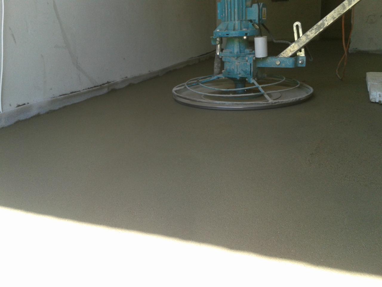 Wygładzona, szara wylewka podłogowa w pomieszczeniu, zacierana mechanicznie zacieraczką. Widoczna ściana i fragment kabla.