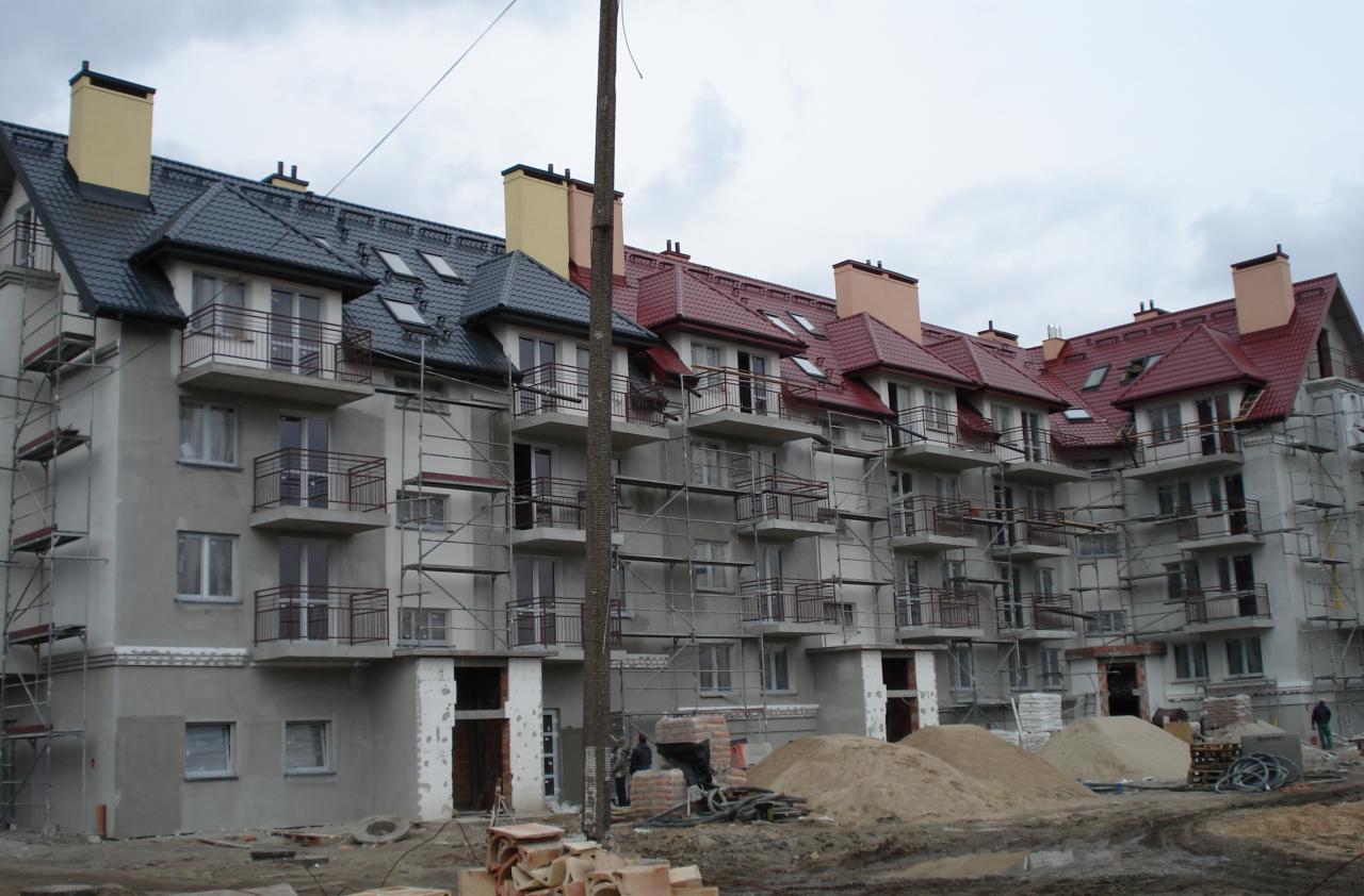 Wielorodzinny budynek w trakcie prac elewacyjnych, z widocznymi rusztowaniami i fragmentami otynkowanej fasady, dachy w dwóch kolorach, na dole hałdy piasku.