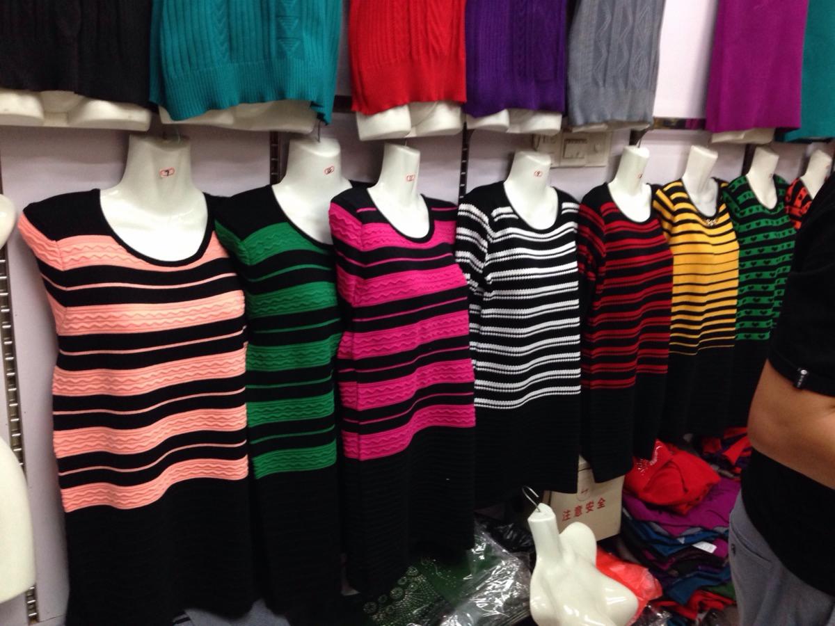 Rząd manekinów prezentujących swetry damskie w paski o różnych kolorach, z widocznymi metkami i stosem ubrań w tle.