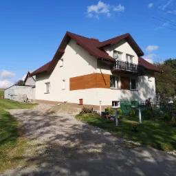 Dwupiętrowy dom z beżową elewacją, brązowym dachem i balkonem z czarną balustradą, widoczny z perspektywy podjazdu w słoneczny dzień. Część elewacji wykończona drewnopodobną okładziną.