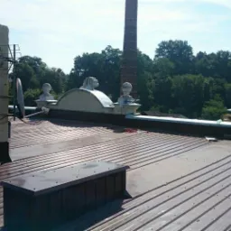 Metalowy dach z kominem i wentylacją na tle zieleni, widok z góry, blacha trapezowa, elementy obróbek blacharskich, komin z anteną satelitarną.