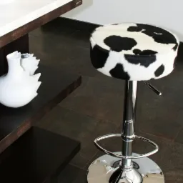 Chromowany hoker barowy z siedziskiem obitym tkaniną we wzór krowich łat, stojący na brązowej podłodze, obok półki z białą wazą.