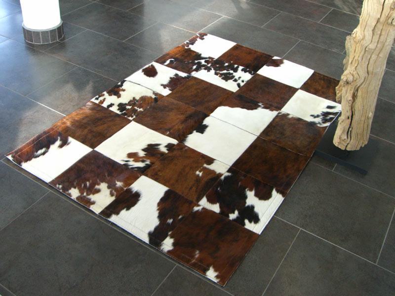 Kwadratowy dywan z brązowo-białych łat imitujących krowią skórę, leżący na ciemnej, płytkowej podłodze obok dekoracyjnego pnia drzewa.