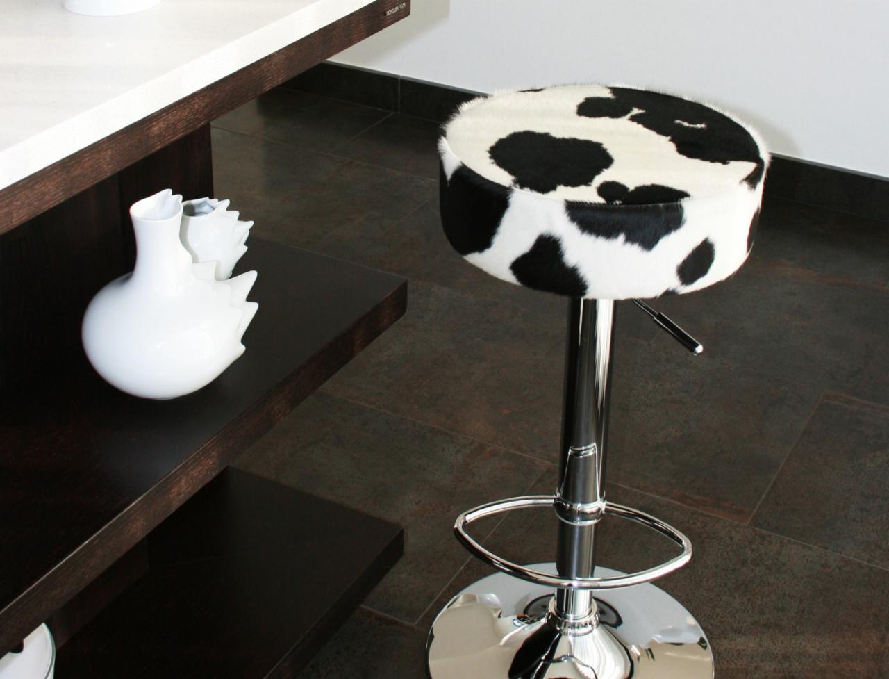 Chromowany hoker barowy z siedziskiem obitym tkaniną we wzór krowich łat, stojący na brązowej podłodze, obok półki z białą wazą.