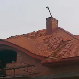 Ceglasty dach z kominem w trakcie krycia, widoczne rusztowania przy niezabudowanym oknie. Częściowo ułożona dachówka na nowym budynku.