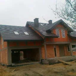 Dom w trakcie budowy z ciemnobrązowym dachem i oknami dachowymi. Widoczna surowa elewacja i elementy konstrukcyjne.  Drzewo bez liści po prawej stronie.