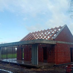 Usługi Remontowo Budowlane Patryk Mierzwa - Surowy stan budowy domu jednorodzinnego z czerwonej cegły, widoczne drewniane krokwie dachu, betonowy wieniec i stalowe podpory, teren wokół placu budowy błotnisty.