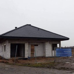 Usługi Remontowo Budowlane Patryk Mierzwa - Surowy dom jednorodzinny z czarnym dachem z dachówki ceramicznej, w stanie deweloperskim, z widocznymi bloczkami betonu komórkowego, na błotnistym terenie, z tablicą reklamową firmy budowlanej w tle.