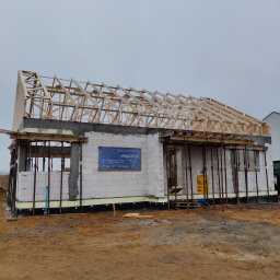 Usługi Remontowo Budowlane Patryk Mierzwa - Surowy stan budynku jednorodzinnego w trakcie budowy: ściany z bloczków, drewniana konstrukcja dachu bez pokrycia, podpory stalowe, tablica informacyjna budowy na ścianie.