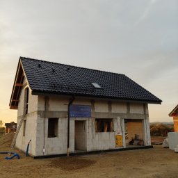 Usługi Remontowo Budowlane Patryk Mierzwa - Surowy stan domu jednorodzinnego w trakcie budowy w miejscowości Biała, z widocznym czarnym dachem z dachówki, ścianami z białego bloczka i zamontowaną rynną. Na ścianie wisi tablica informacyjna...